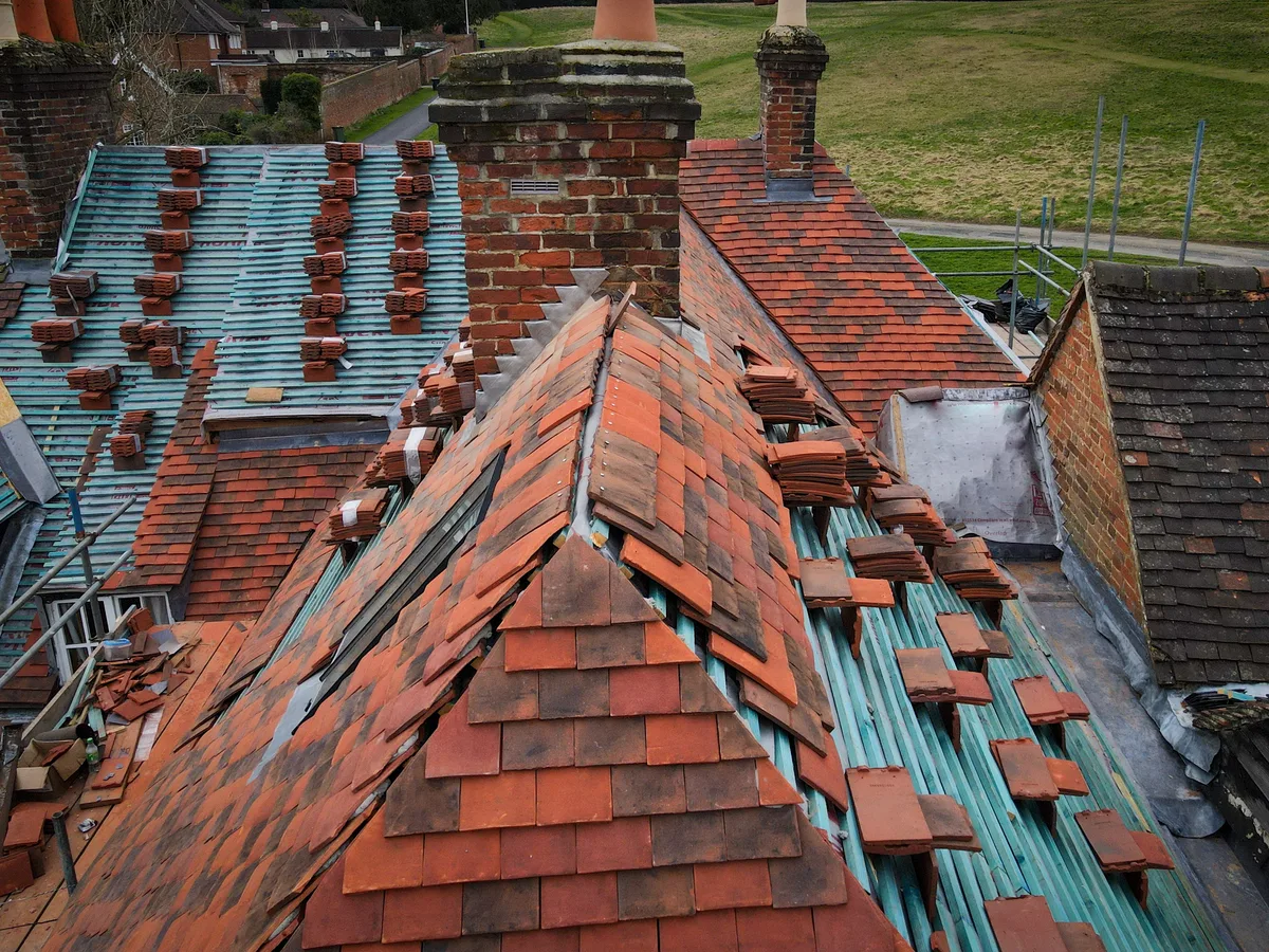 An old clay tile roof around a chimney, partly stripped with loose tiles stacked on the battens
